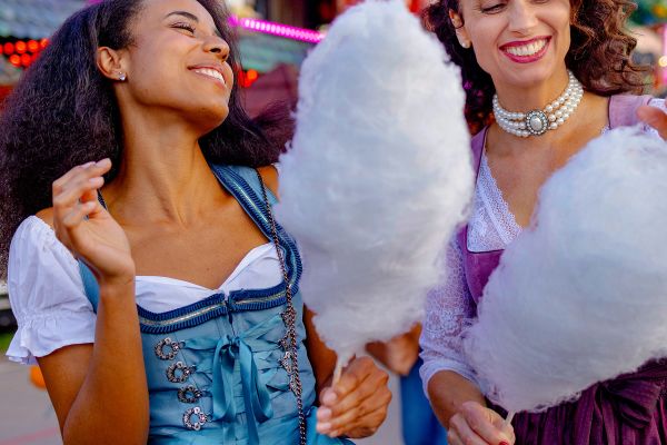 Two female best friends in traditional Bavarian Dirndl dresses eating cotton candy while walking at a beer festival in Munich.