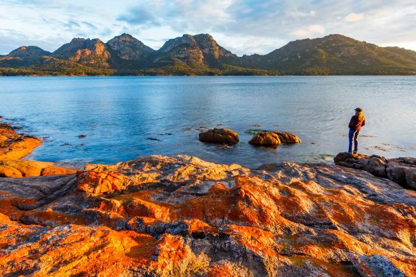 A lady standing on rocks next to a lake with mountains in the distance