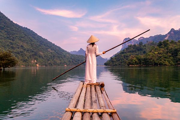 Person in a white dress and straw hat, holding a long staff while standing on a bamboo raft in a river