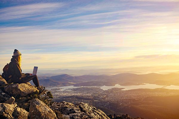 Woman sitting on a rock with a laptop looking out at Hobart at sunset