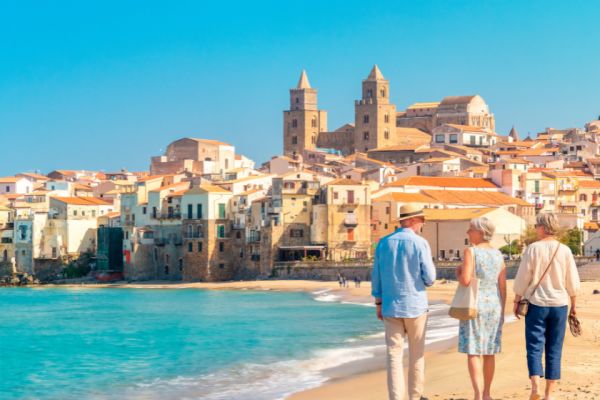 Three travellers strolling along a sunlit beach beside a historic Mediterranean seaside town with turquoise waters and relaxed coastal charm