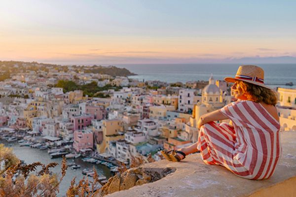 Woman in a stripped dress sitting on a wall overlooking a coastal city at sunset