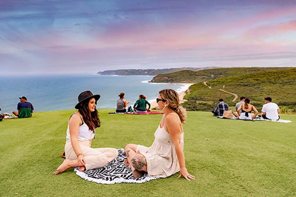 Couples on picnic blankets on a grassy hill, looking out at the sea at sunset