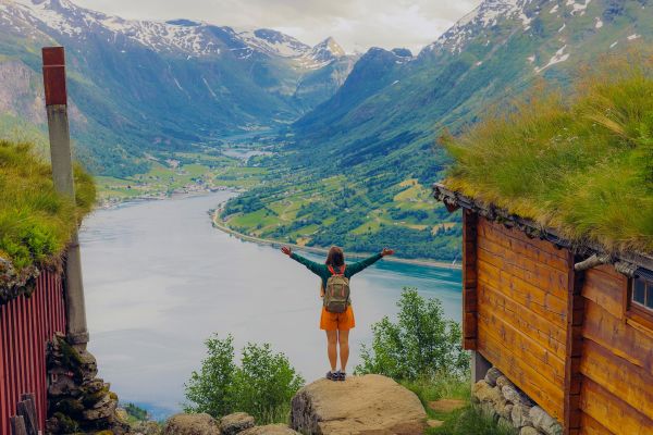 Person standing between two wooden buildings with grass growing on the roofs, looking out at a Fjord
