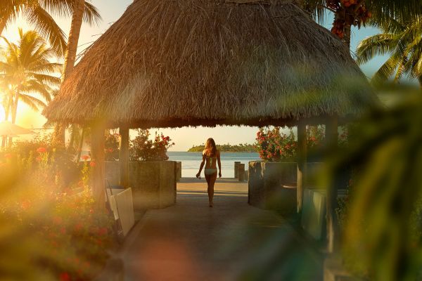 Wide shot of a woman in a white bikini under a thatched roofed pagoda looking at the ocean