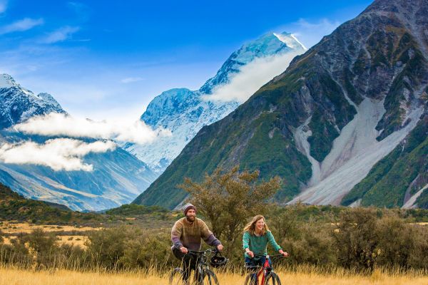 Couple riding bikes through grassy fields with snow-covered mountains in the background