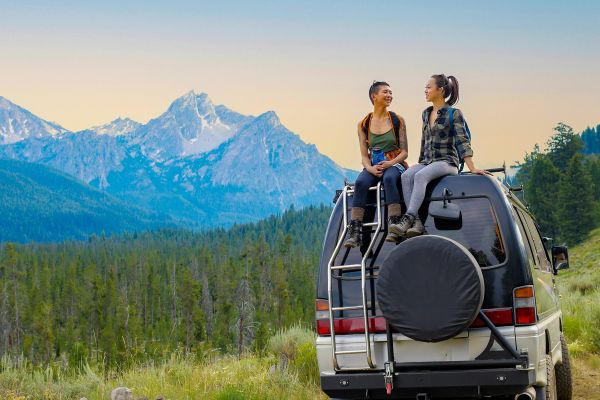 Couple sitting on the roof of their car, with hills and pine forest in the background