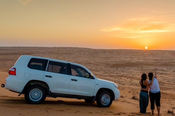 Couple hugging next to their white car, looking at a sunset over a large sand dune