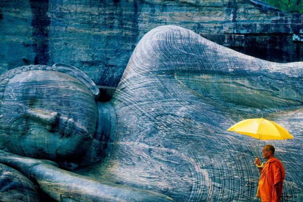 Monk holding a yellow umbrella looking at a fallen Buddha statue