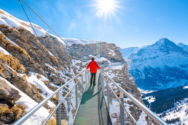Person on a thin suspensions bridge looking out at snowy mountains on a sunny day