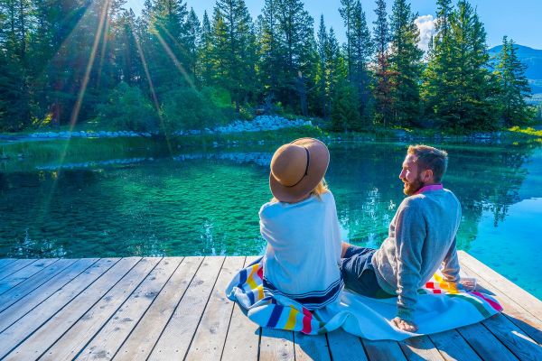Couple sitting on a pier looking at a crystal clear lake with tall trees on the opposite shore