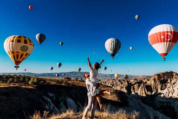 Man lifting a woman into the air on a grassy hill while dozens of hot air balloons take off around them