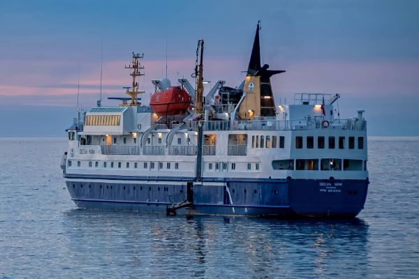 The expedition cruise ship Ocean Nova, painted white and blue, anchored on calm water at dusk, with a soft pink and blue sunset sky reflected on the still sea surface.