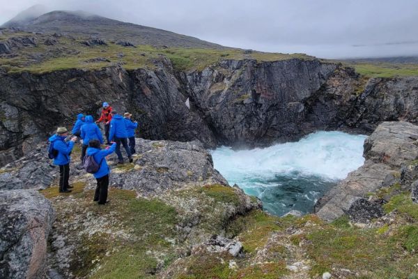 A group of travellers in matching blue jackets and one red jacket stand at the edge of a dramatic rocky cliff, looking down into a churning turquoise sea surge pool carved into the rugged coastline. A moss-covered mountain rises in the background under an overcast sky.