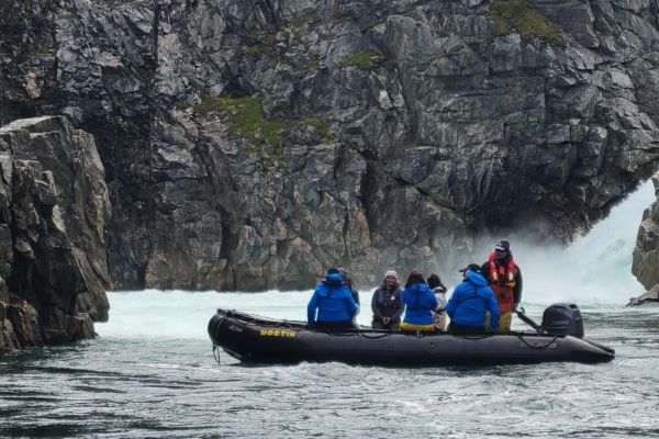 A small group of passengers in blue expedition jackets sit aboard a black inflatable Zodiac boat labelled "Bootis," approaching a powerful waterfall cascading through a narrow gap in towering dark rock cliffs, with white water churning below.