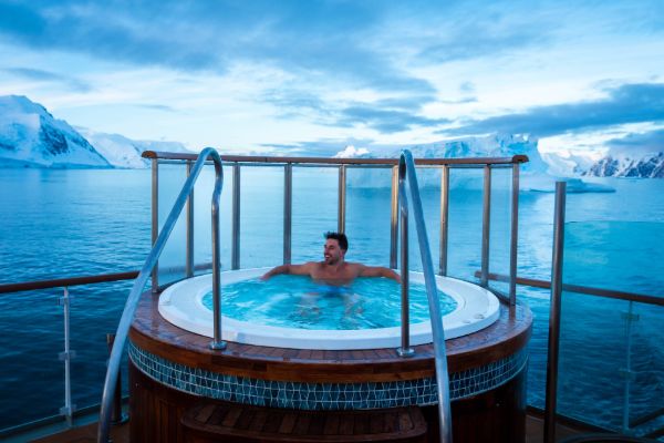 A man relaxes alone in a circular outdoor hot tub on the deck of an expedition cruise ship, looking out over a calm polar sea dotted with icebergs and snow-covered mountains under a moody blue twilight sky.
