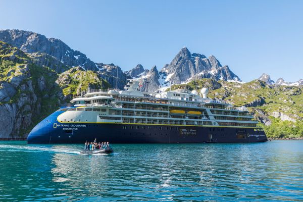 The National Geographic Endurance expedition cruise ship anchored in a stunning fjord with jagged, snow-dusted mountain peaks rising sharply behind it under a clear blue sky. A small inflatable Zodiac boat with passengers heads back toward the ship across the calm, deep-blue water.
