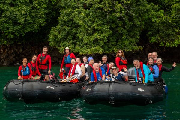 A large, smiling group of around eighteen expedition passengers and crew in red and blue life vests pose cheerfully across two black inflatable Zodiac boats on calm green water, with lush tropical jungle vegetation visible behind them.