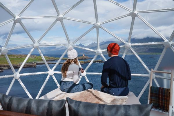 Two people wearing woolly hats sit side by side inside a transparent geodesic dome on the deck of an expedition ship, looking out over a calm coastal waterway with a small island, lighthouse, and distant mountain ranges visible through the dome's white framework under a partly cloudy sky.