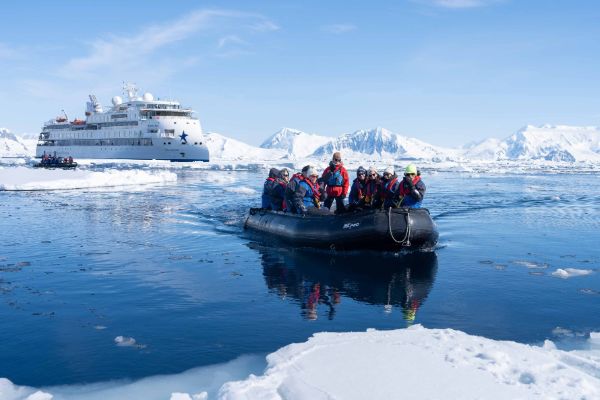 A group of expedition passengers in colourful cold-weather gear ride a black inflatable Zodiac boat through icy, partially frozen Antarctic waters, with a small white expedition cruise ship anchored among snow-covered mountains and ice floes in the background under a clear blue sky.