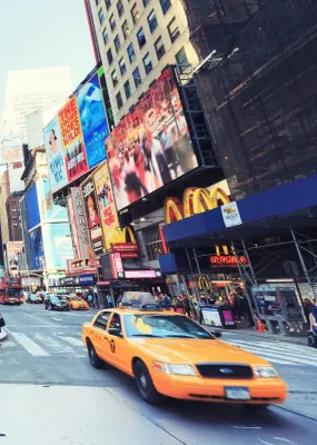 A busy crosswalk in Times Square with people crossing the road and cars driving down the street.