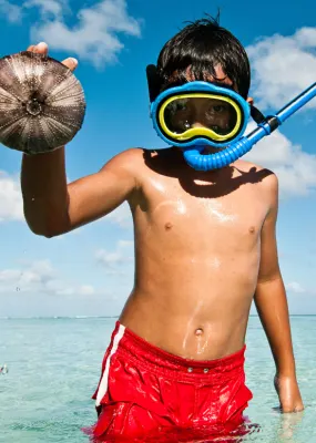 A boy wearing  a mask and snorkel stands in a lagoon holding up a sea urchin