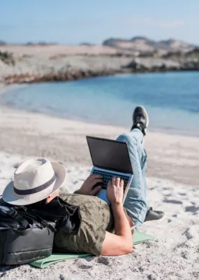 Man lying on beach resting head on backpack while working on his laptop