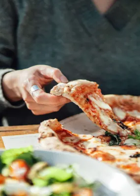 Lady wearing grey sweater lifting the first piece of pizza from board on table at restaurant