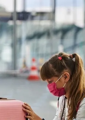 Woman wearing a mask adjusting a child's mask in an airport while another child wearing a mask sits next to her suitcase