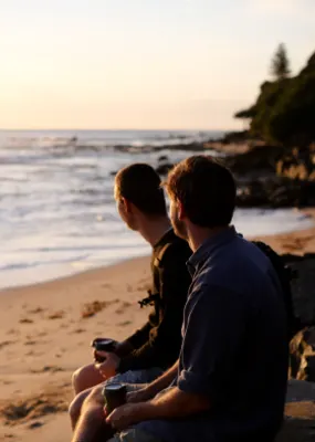 Two gentlemen sitting on a rock enjoying a cup of coffee while gazing out into the sea