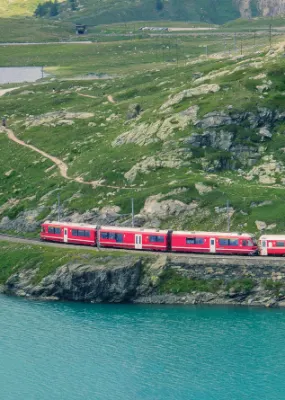 Red train going past a lake and green mountains