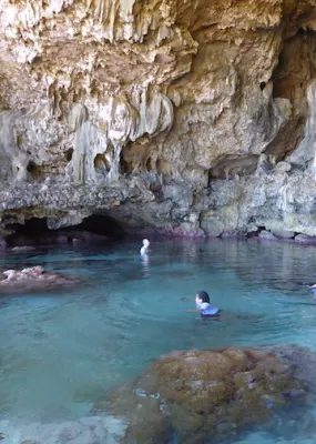 People swim in a large ocean pool in a limestone cavern with high pocked walls where other people can be seen climbing.