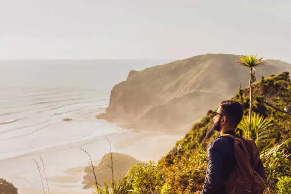 Bearded man wearing a backpack staring out at the ocean from atop a grassy cliff