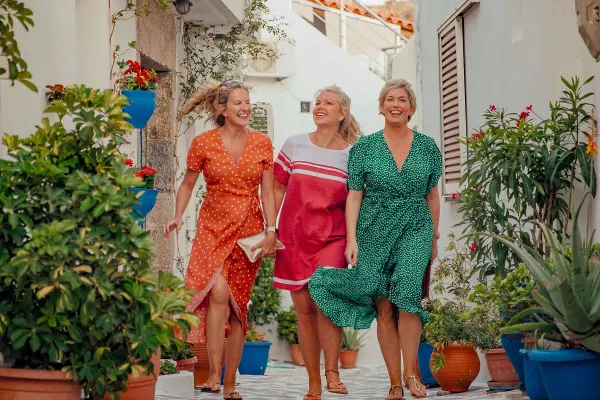 Three women in flowing dresses walking through a street lined by potted plants