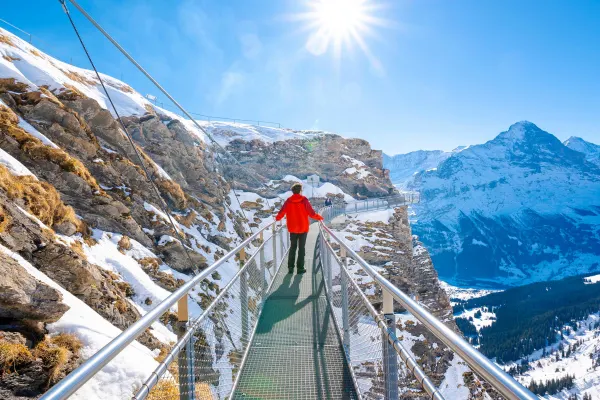 Person on a thin suspensions bridge looking out at snowy mountains on a sunny day