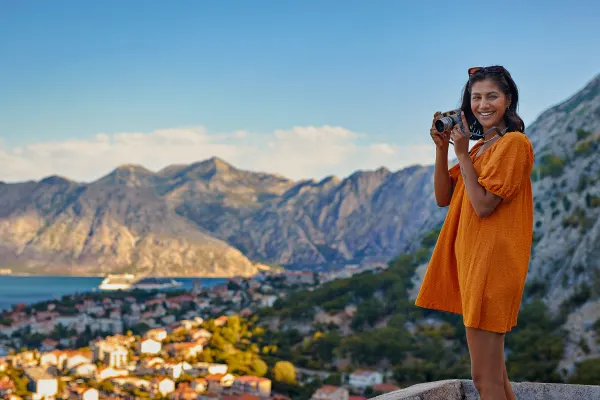 Woman in an orange dress holding a camera, with a coastal town and mountains in the background