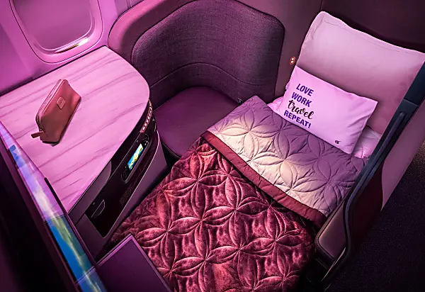 Qatar Airways clean business class seat with two pillows and a quilt on the reclined bed and a complementary toiletries package on the side table.