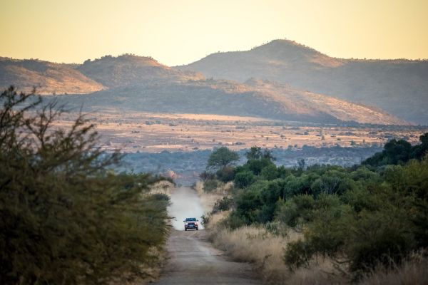 A car driving on dirt road in between bush land with sunset and mountains in background