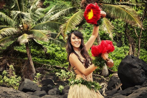 Attractive young hula dancer, dancing on a Hawaiian beach with red feathered shakers ('Uli 'Uli) and palm trees in the background.