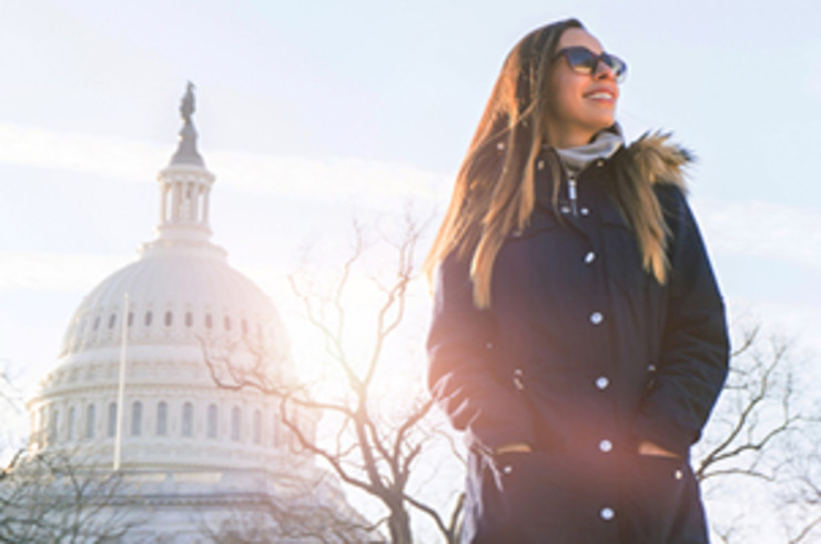 A person standing outdoors near the United States Capitol in Washington, D.C. They’re dressed warmly in a dark winter coat with a fur hood and wearing sunglasses, suggesting chilly weather. The iconic Capitol dome rises prominently in the background, partially lit by sunlight