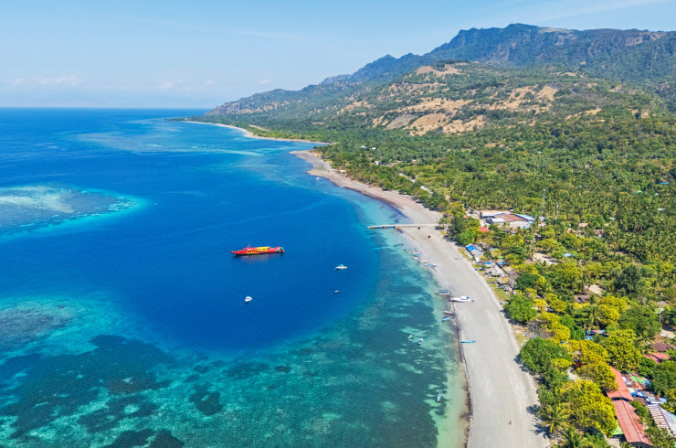 Aerial view of East Timor's coastline with vivid blue waters lapping a palm-fringed beach beneath rugged mountain hills
