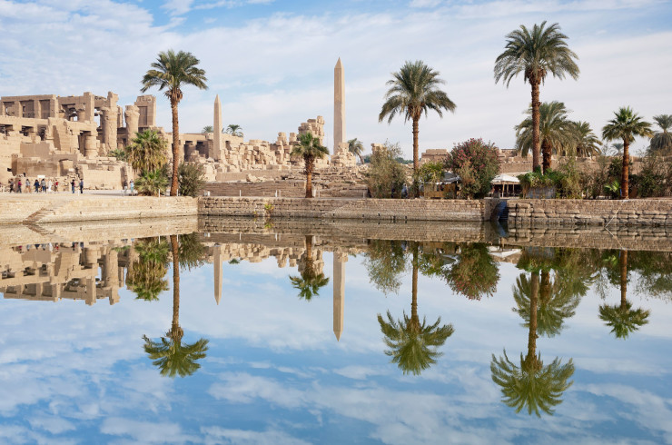 Egyptian obelisks and sandstone ruins lined by palm trees and a clear lake