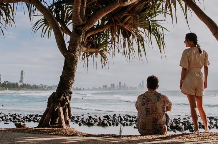 Couple shaded by large beach trees sitting on the beach looking out at skyscrapers on the coastline