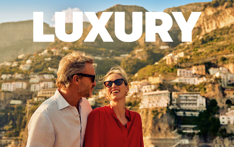 Two middle aged people smiling, sailing past hotels and apartments on steep cliffs