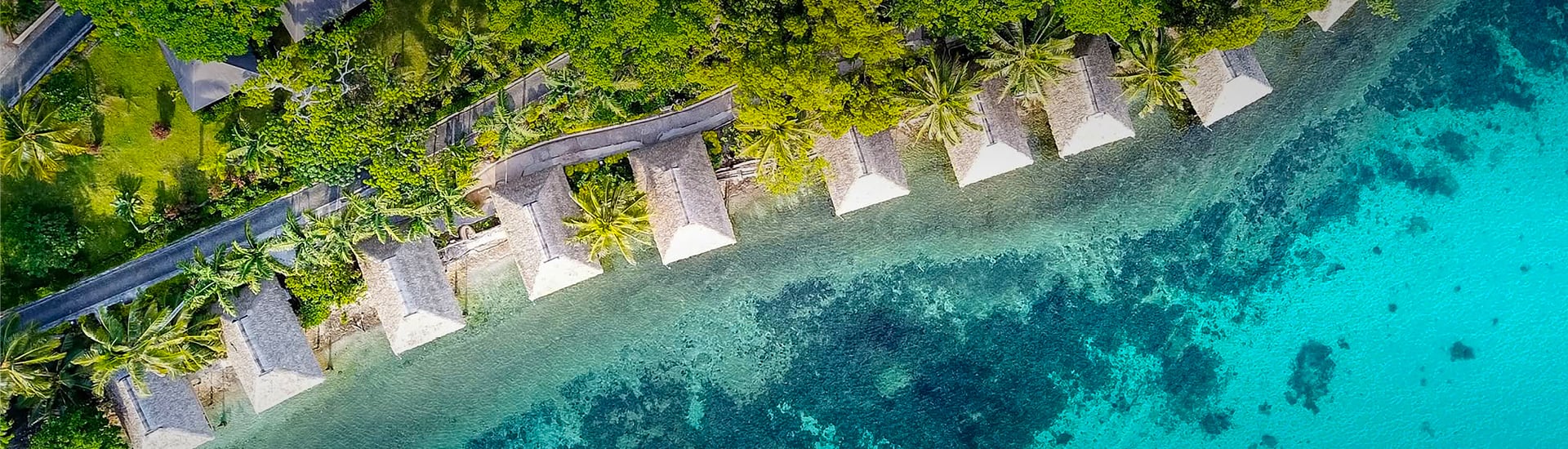 Aerial view of overwater bungalows nestled among tropical palms along a turquoise coral reef shoreline on a Pacific island