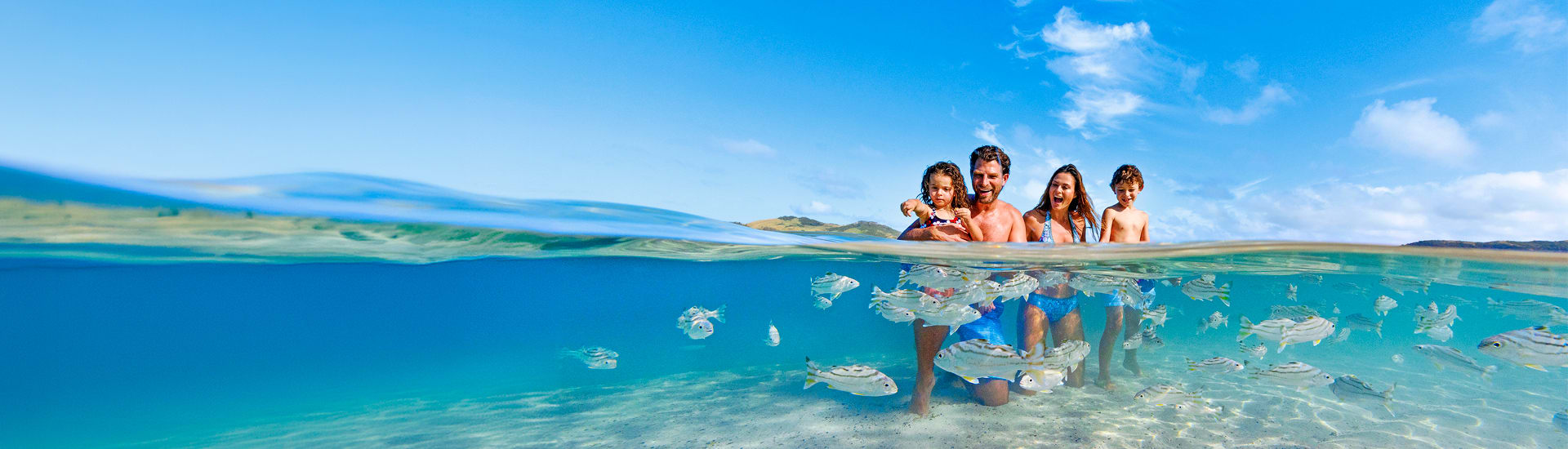 Smiling family wading together through clear turquoise shallows with fish swimming around them for family holidays where kids stay and eat free