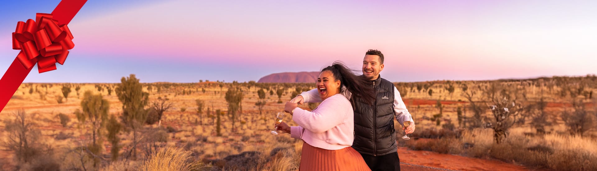couple dancing out the front of uluru at sunset with a red bow in the corner of the image 