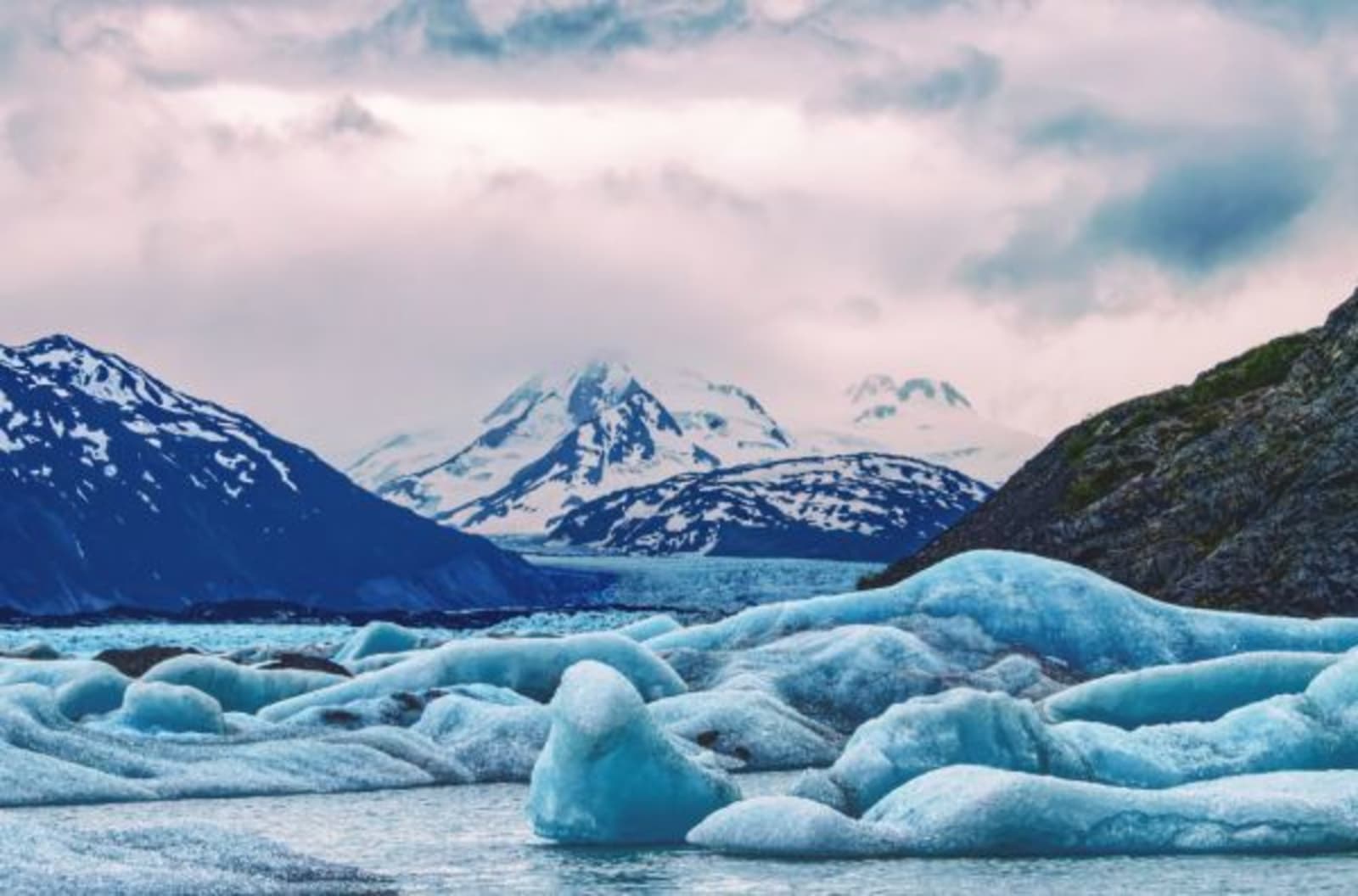 Glacier with a snow-capped mountain in the background and a cloudy sky.