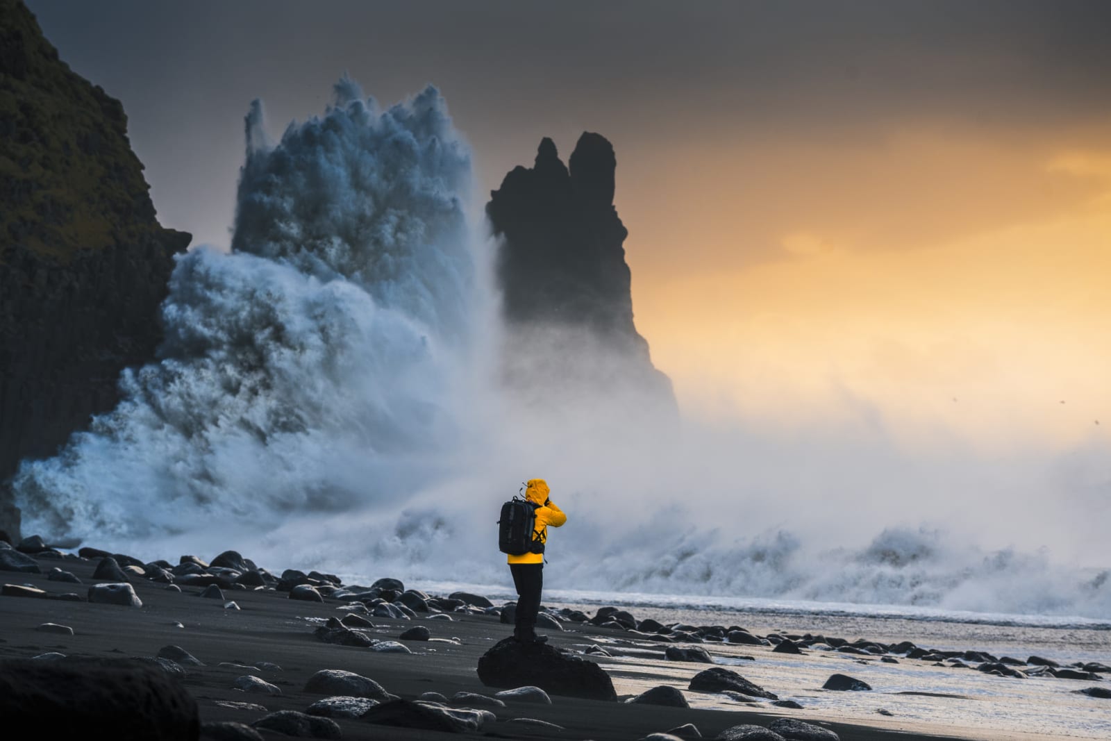 Explorer stands on rocky shore with dramatic waves against sunset backdrop in Iceland