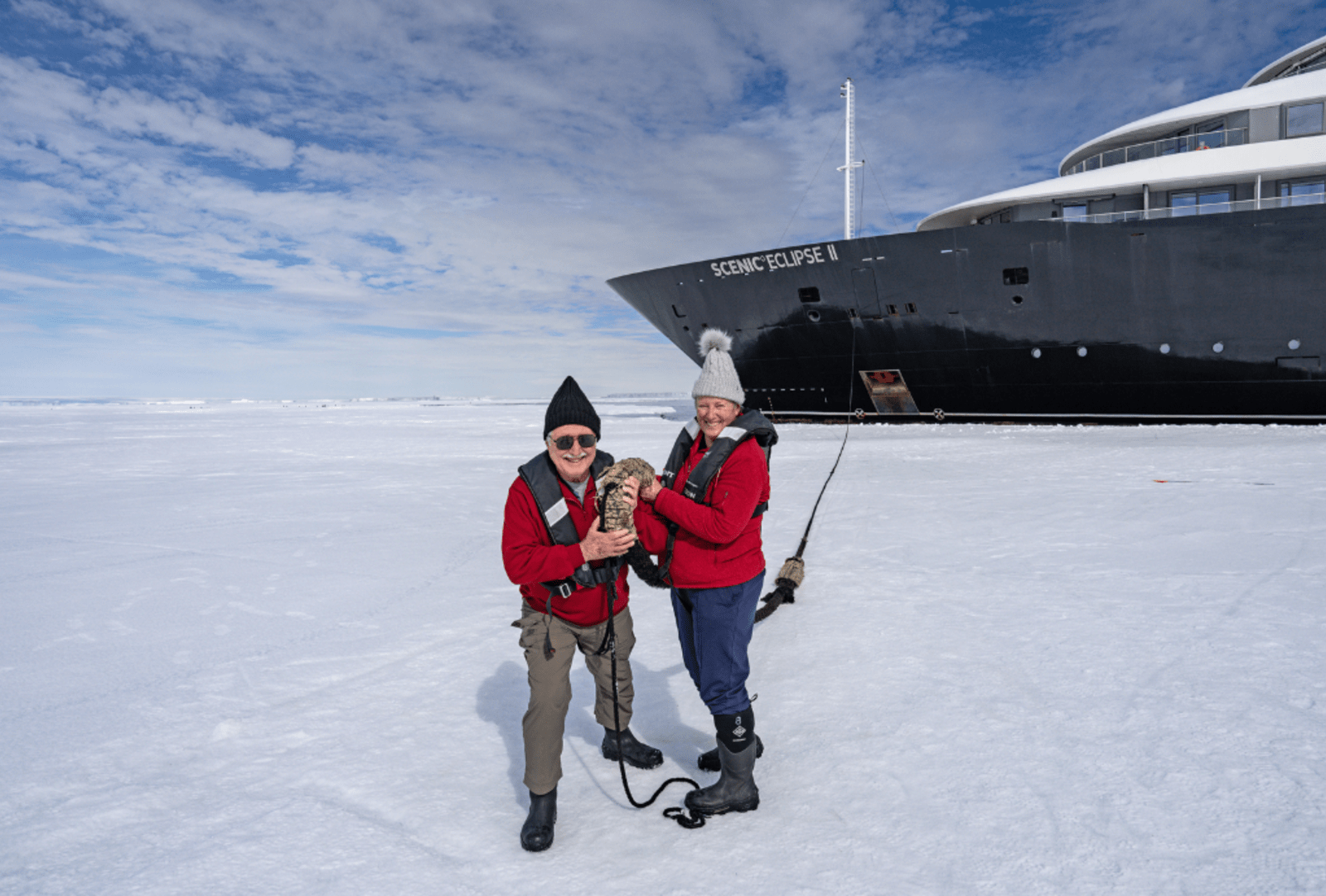 Couple bundled in red jackets holding whale bone on the ice beside the Scenic Eclipse II ship